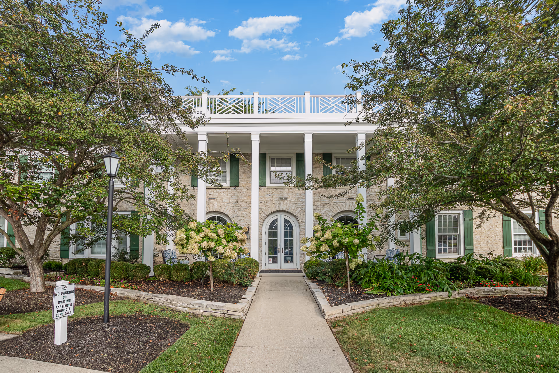Front entrance of a two-story stone building with white columns, green shutters, a central arched doorway, and landscaped walkway framed by trees.