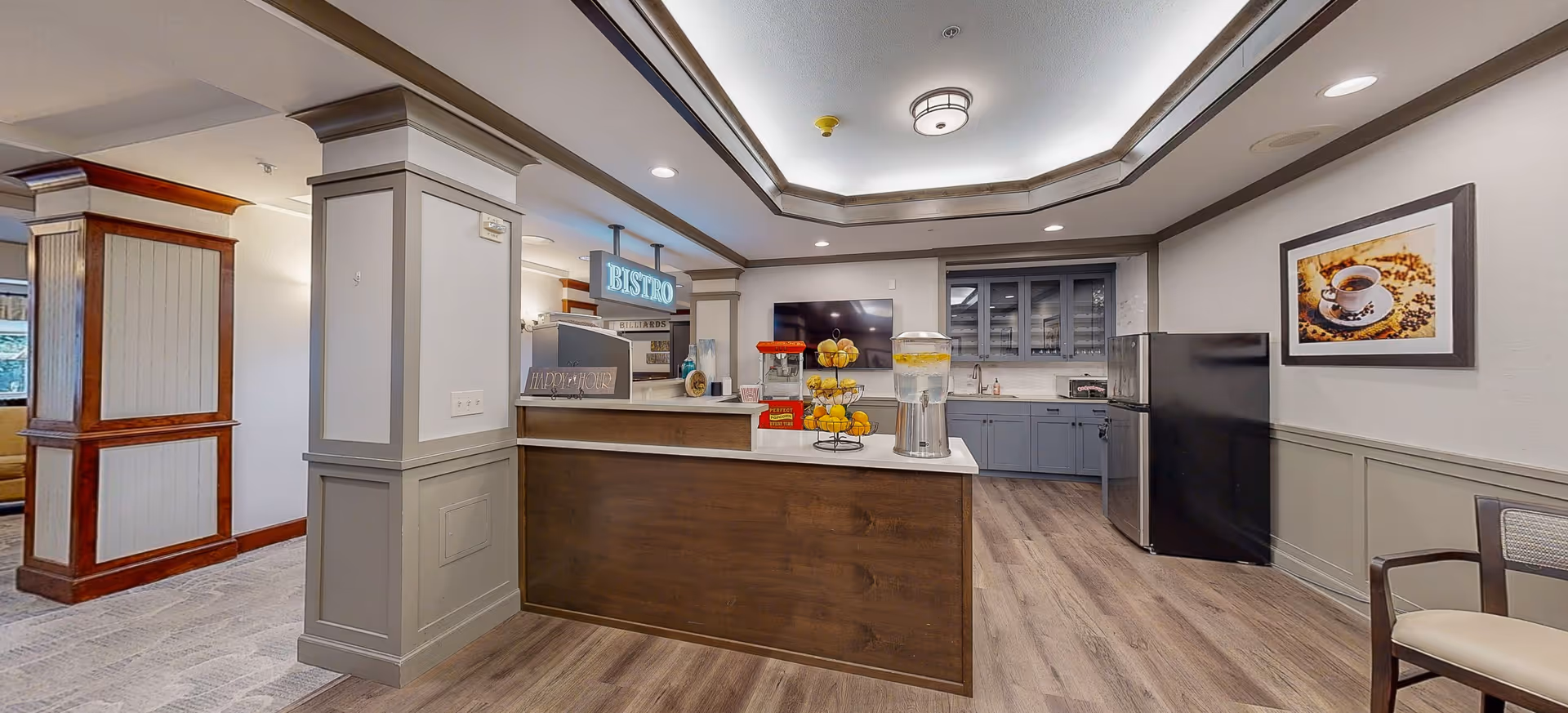 Interior view of a senior living facility's bistro area with a counter displaying a popcorn machine, a tiered fruit stand, and a beverage dispenser. Behind the counter are cabinets, a refrigerator, and a wall-mounted TV. The space features wood and gray tones with a framed picture of a coffee cup on the wall and a chair to the right.