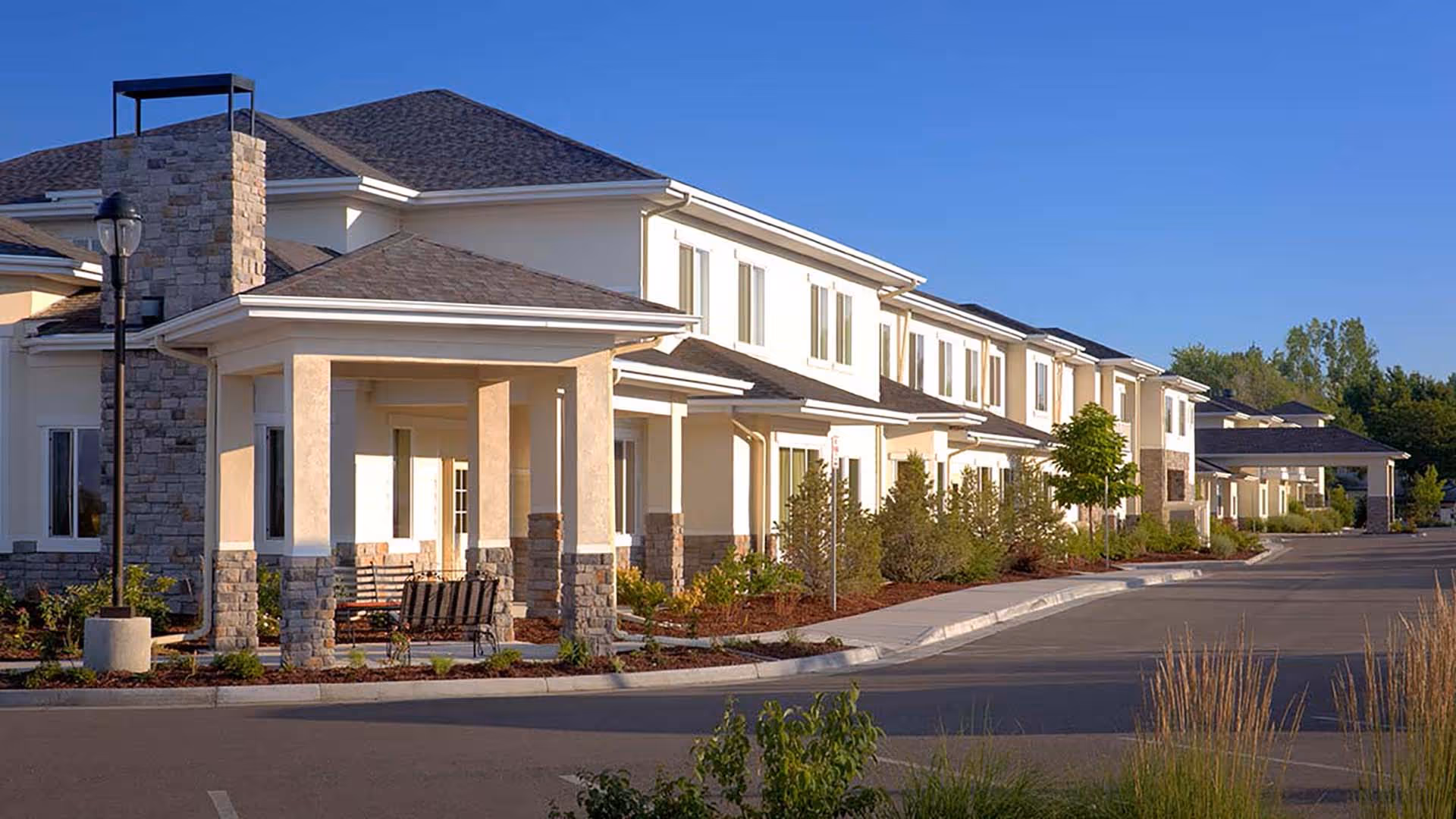 Exterior view of a senior living facility building with beige walls, stone accents, multiple windows, and a covered entrance area with benches. The facility is surrounded by landscaped greenery and a paved driveway under a clear blue sky.