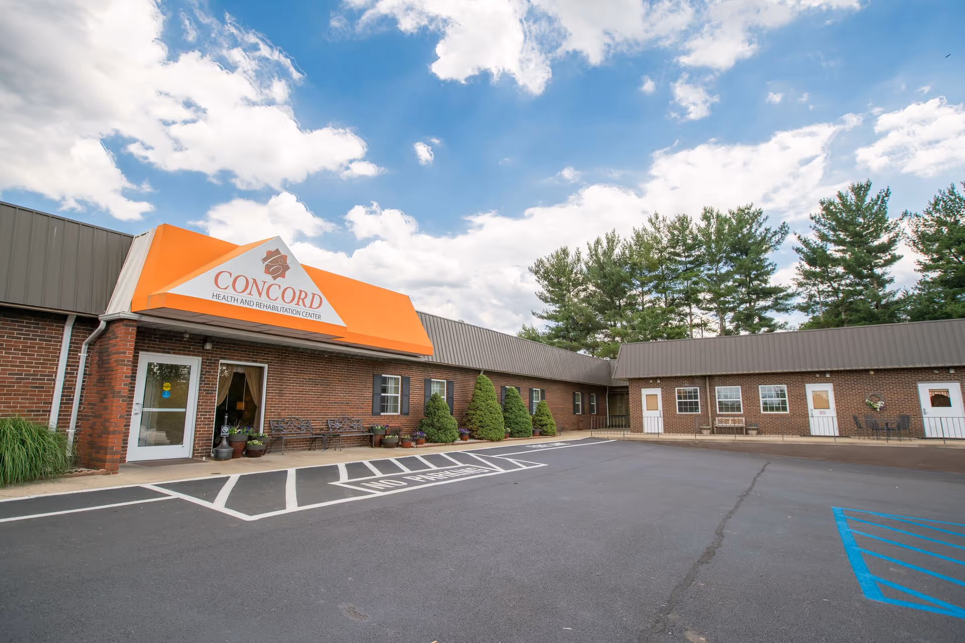 Exterior view of Concord Health and Rehabilitation Center building with a brick facade, an orange awning above the entrance, several windows, doors, benches, and some greenery. The parking lot in front has marked spaces including a handicapped spot. Trees and a partly cloudy sky are visible in the background.