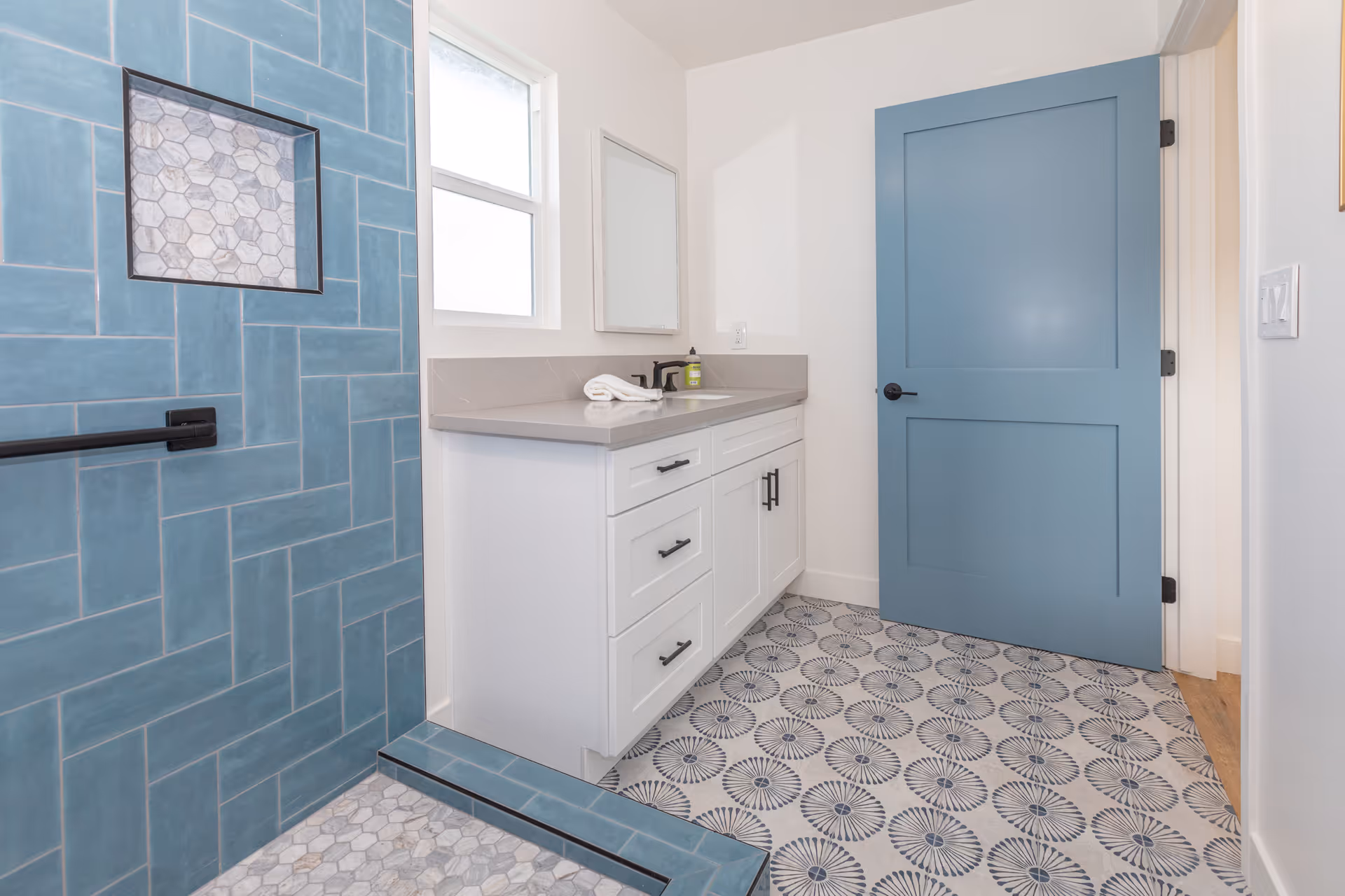 A modern bathroom featuring a blue tiled shower with a built-in niche, a white vanity with a gray countertop, a rectangular mirror above the sink, a blue door, and patterned floor tiles in blue and white.