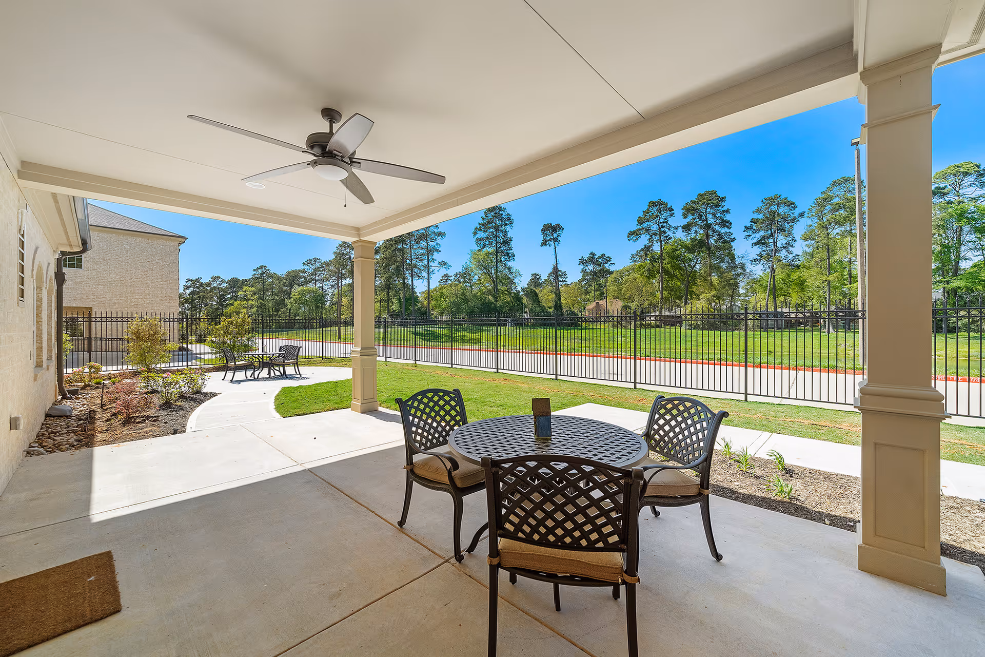 Covered outdoor patio area with a ceiling fan, a round metal table with four cushioned chairs, and a view of a fenced grassy yard with trees in the background under a clear blue sky.
