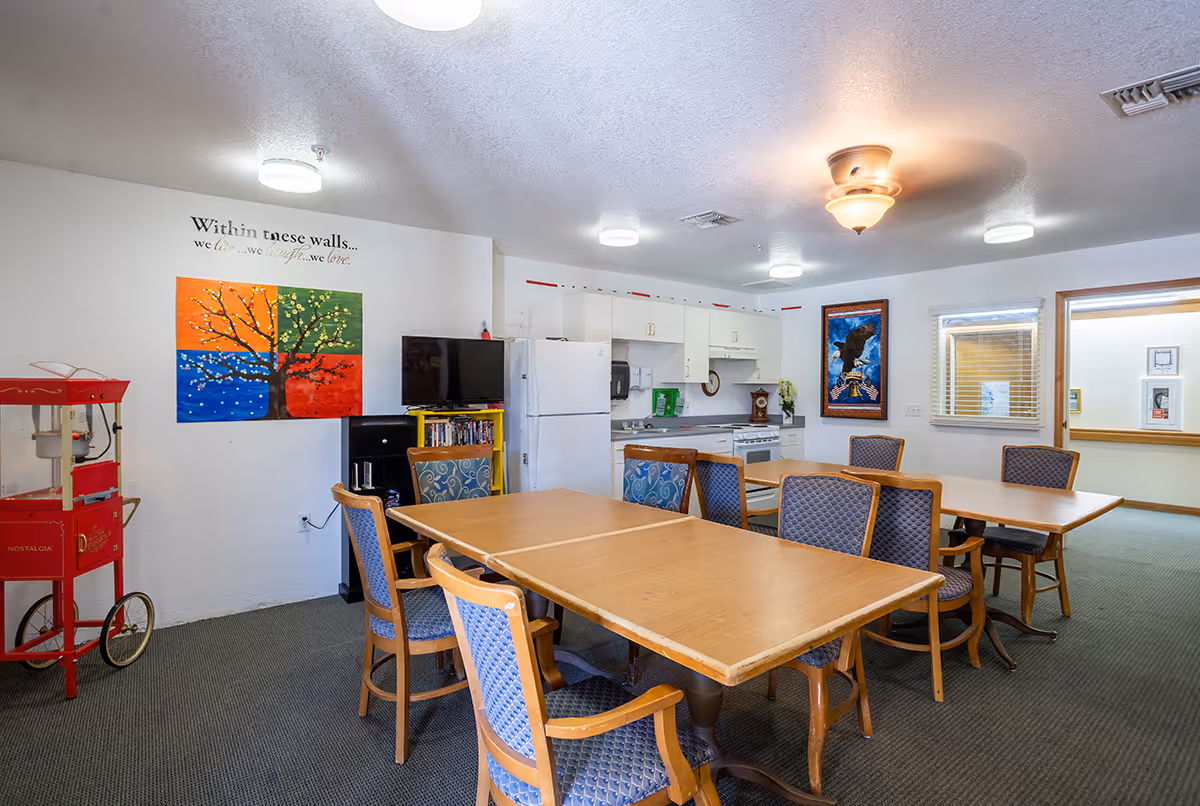 A communal dining and kitchen area in a senior living facility featuring two wooden tables with blue cushioned chairs, a white refrigerator, stove, microwave, and cabinets. On the left side, there is a red popcorn machine and a colorful painting of a tree with four quadrants in orange, green, blue, and red. Above the painting, a wall decal reads 'Within these walls... we live... we laugh... we love.' A TV and bookshelf are also visible, along with a framed eagle picture on the far wall.