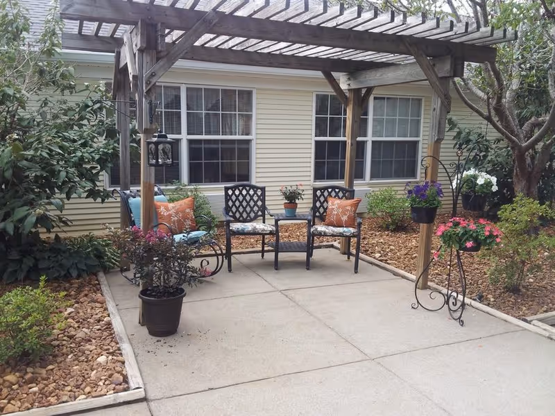 Outdoor patio under a wooden pergola with chairs, cushions, and potted flowers in front of a building with windows.