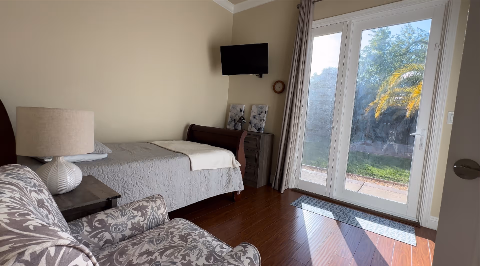 A cozy bedroom with a single bed covered in a gray quilt, a patterned armchair, a bedside table with a lamp, a small dresser with decorative items, a wall-mounted TV, and large glass doors leading to an outdoor area with greenery and a palm tree.