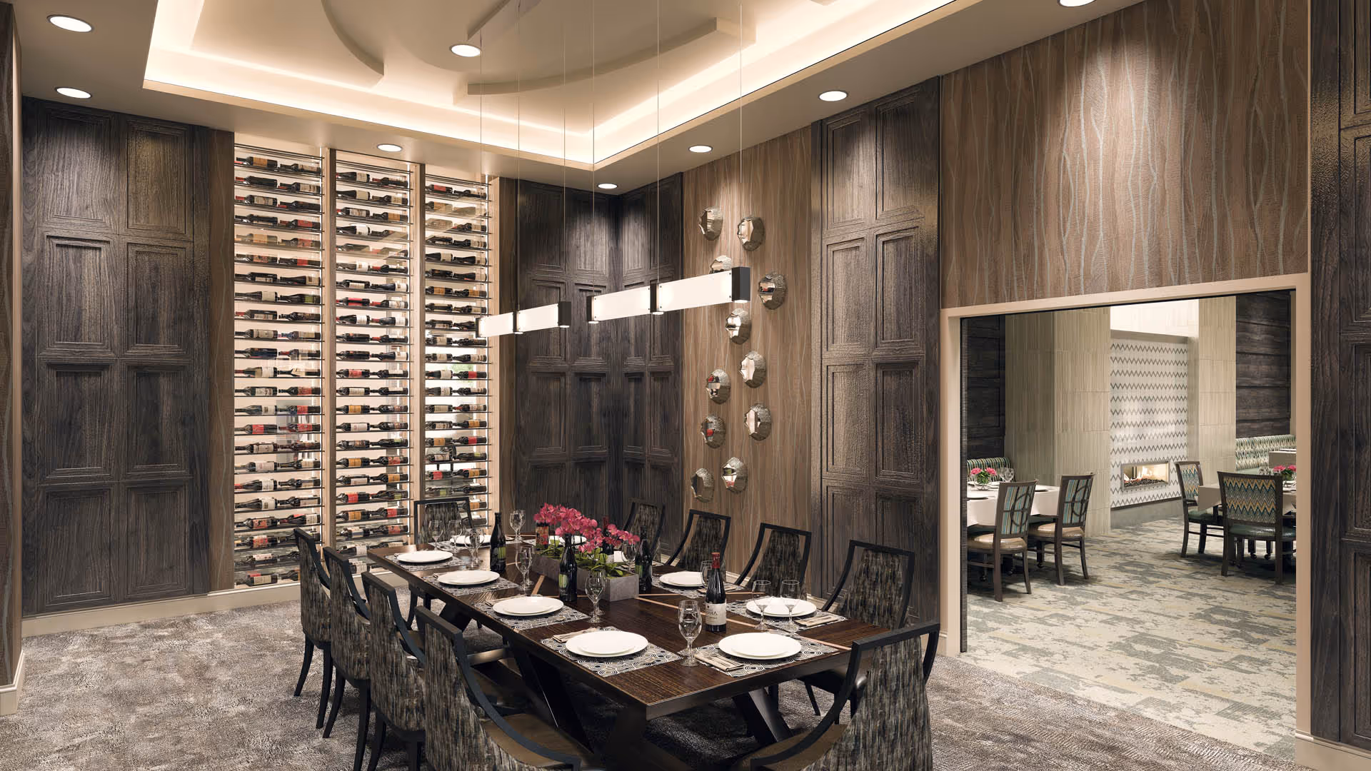 A modern dining room with a long rectangular table set for eight people, featuring plates, wine glasses, and a bottle of wine. The room has dark wood paneling on the walls, a large wine rack filled with bottles, and contemporary pendant lights hanging from the ceiling. Through an open doorway, another dining area with a fireplace and additional tables and chairs is visible.