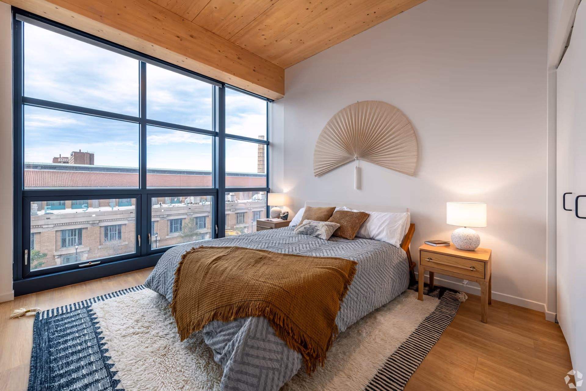 A modern bedroom with a large window showing an urban view. The room features a bed with gray bedding and a brown throw blanket, two wooden nightstands with lamps, a decorative fan on the wall above the bed, and a patterned rug on the wooden floor.