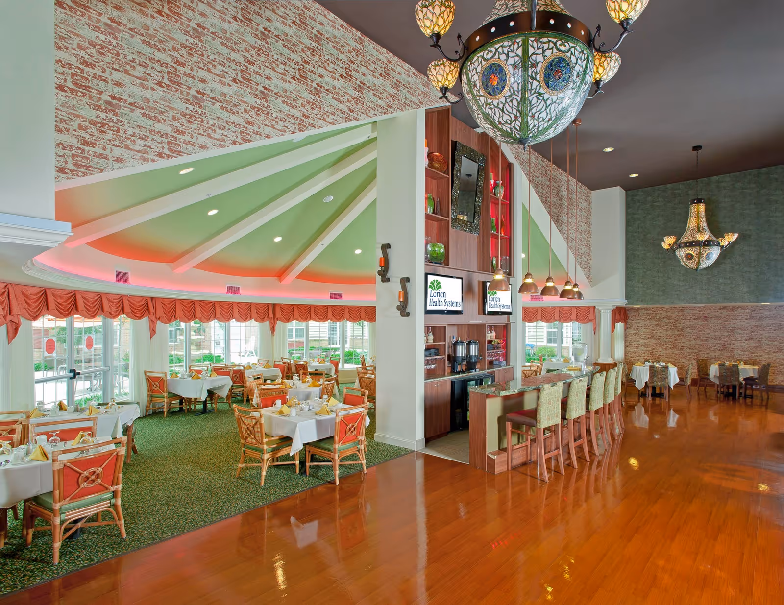 A spacious dining area with tables covered in white tablecloths and set with napkins and glassware. The room features a green carpeted section with large windows and red valances, and a polished wooden floor section with a bar counter and high chairs. Decorative chandeliers hang from the ceiling, and there are two screens displaying the Lorien Health Systems logo.