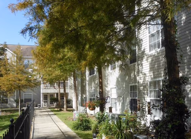 Outdoor walkway alongside a white multi-story senior living community building with trees, plants, and a black metal fence on the left side.