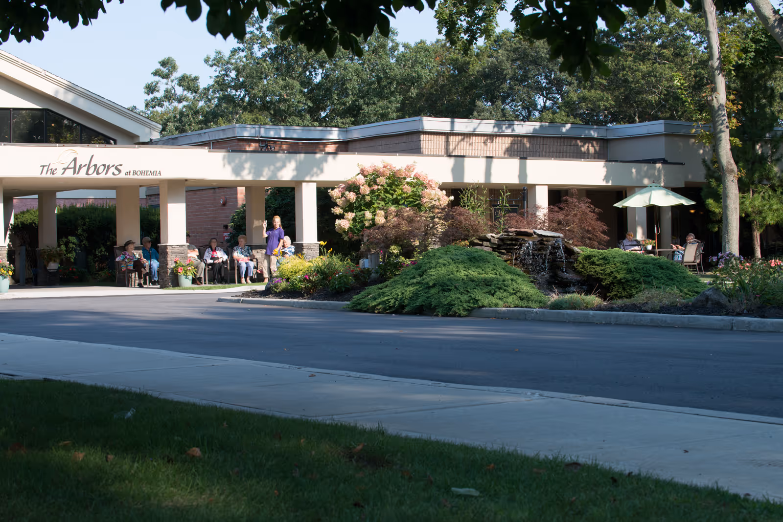 Front entrance of The Arbors senior living facility with a covered driveway, seated residents near columns, and landscaped shrubs and a small fountain.