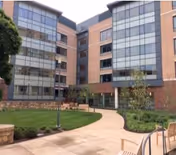 Outdoor courtyard area of a senior living facility with a curved paved walkway, green lawn, benches, small trees, and a multi-story building with large windows in the background.