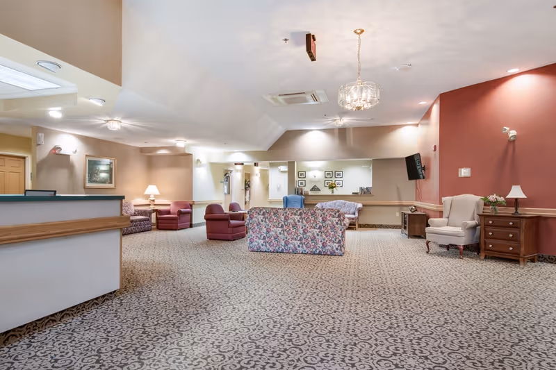 A spacious senior living facility common area with patterned carpet, floral and solid-colored armchairs and sofas arranged for seating. The walls are painted in neutral tones with one accent wall in red. There is a reception desk on the left side, a wall-mounted TV on the right, and decorative lighting fixtures on the ceiling. A small table with a lamp and flowers is also visible.