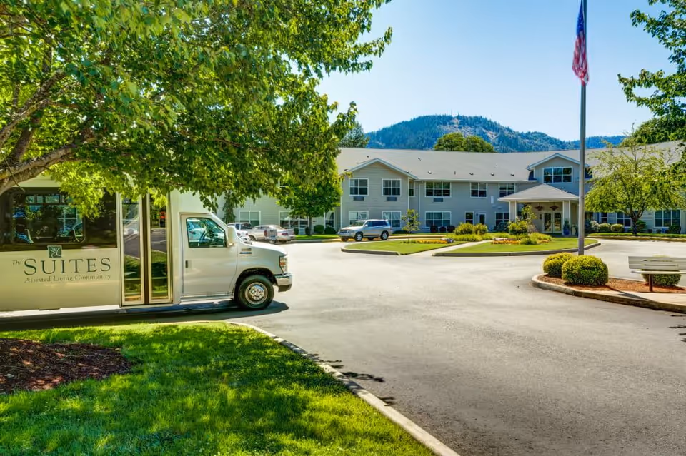 Exterior view of The Suites Assisted Living Community building with a driveway, parked cars, a shuttle van, green trees, and a flagpole with an American flag under a clear blue sky and mountains in the background.