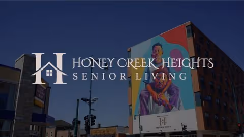 Exterior view of a multi-story building with a large colorful mural of a person on one side under a clear blue sky. The building has the text 'Honey Creek Heights Senior Living' displayed prominently.