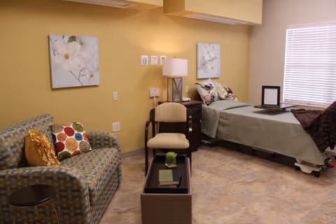 A furnished senior living bedroom with a bed, sofa, chair, side table and wall art.