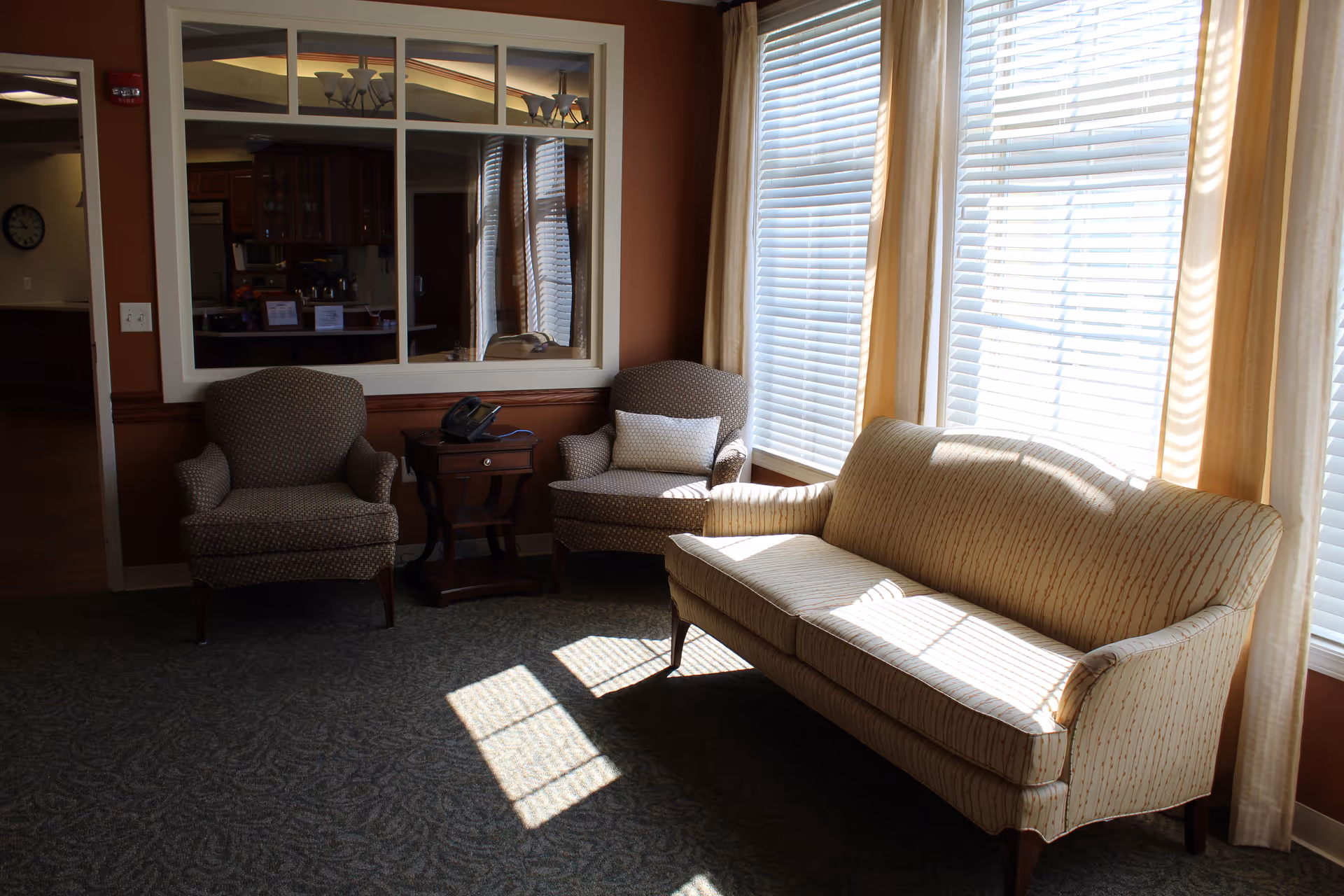 A cozy sitting area with two patterned armchairs and a beige striped sofa placed near large windows with blinds. A small wooden side table with a telephone is positioned between the armchairs. The walls are painted a warm brown color, and there is a large window opening into another room, reflecting a chandelier and kitchen cabinets.