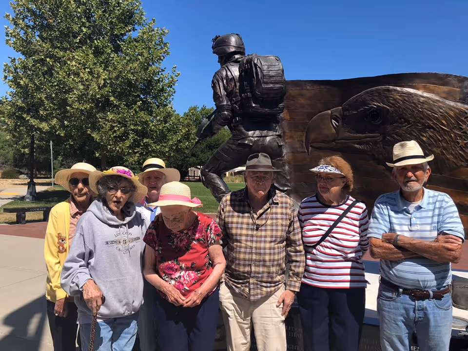 A group of seven elderly people wearing hats standing outdoors in front of a large bronze statue depicting a soldier and an eagle. The sky is clear and blue, and there are green trees in the background.