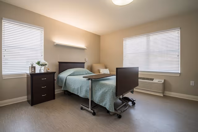 A simple, clean bedroom in a senior living facility featuring a single hospital-style bed with a light blue bedspread, a dark wooden nightstand with a vase of flowers and a lantern, a beige armchair with a decorative pillow, two windows with white blinds, and a wall-mounted air conditioning unit below one window.