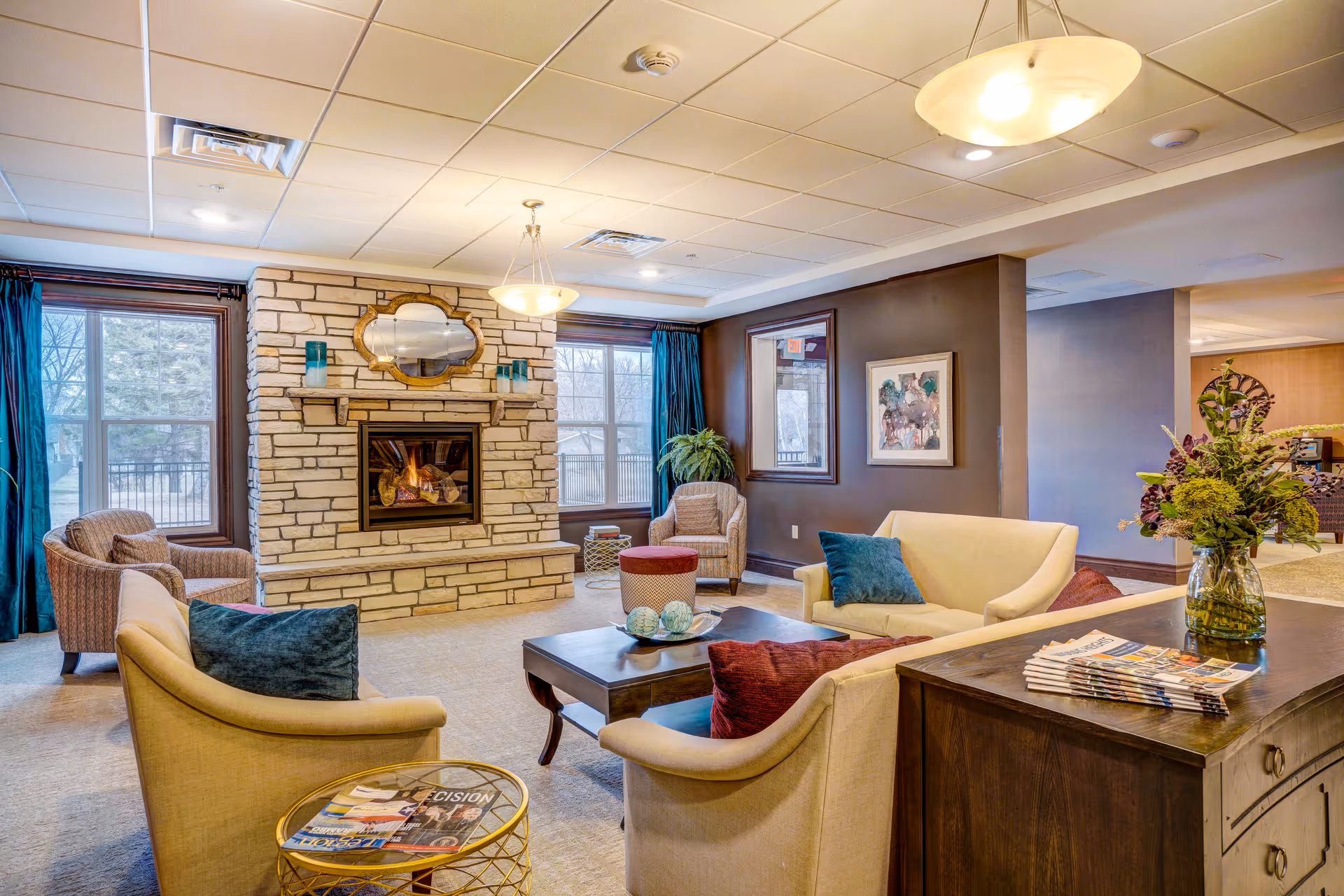 A cozy living room area in a senior living facility featuring a stone fireplace with a mirror above it, two large windows with blue curtains, several armchairs and sofas with colorful cushions arranged around a wooden coffee table, a side table with magazines, and a wooden cabinet with a vase of flowers and more magazines.