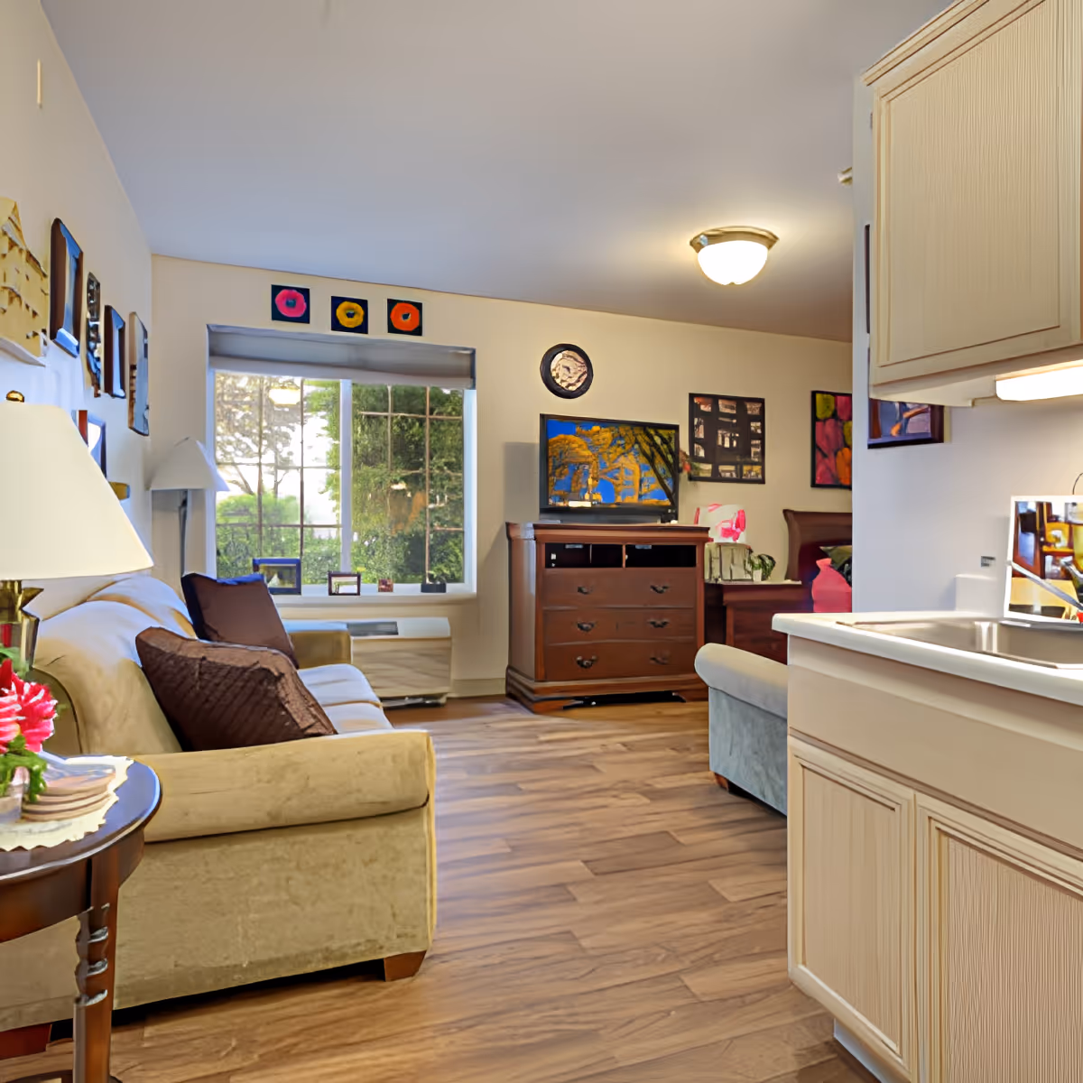 A cozy living room area in an assisted living facility featuring a beige sofa with brown pillows, a wooden side table with a lamp and flowers, a window with a view of greenery outside, a wooden dresser with a TV on top, colorful artwork on the walls, and a partial view of a kitchen sink and cabinets on the right.