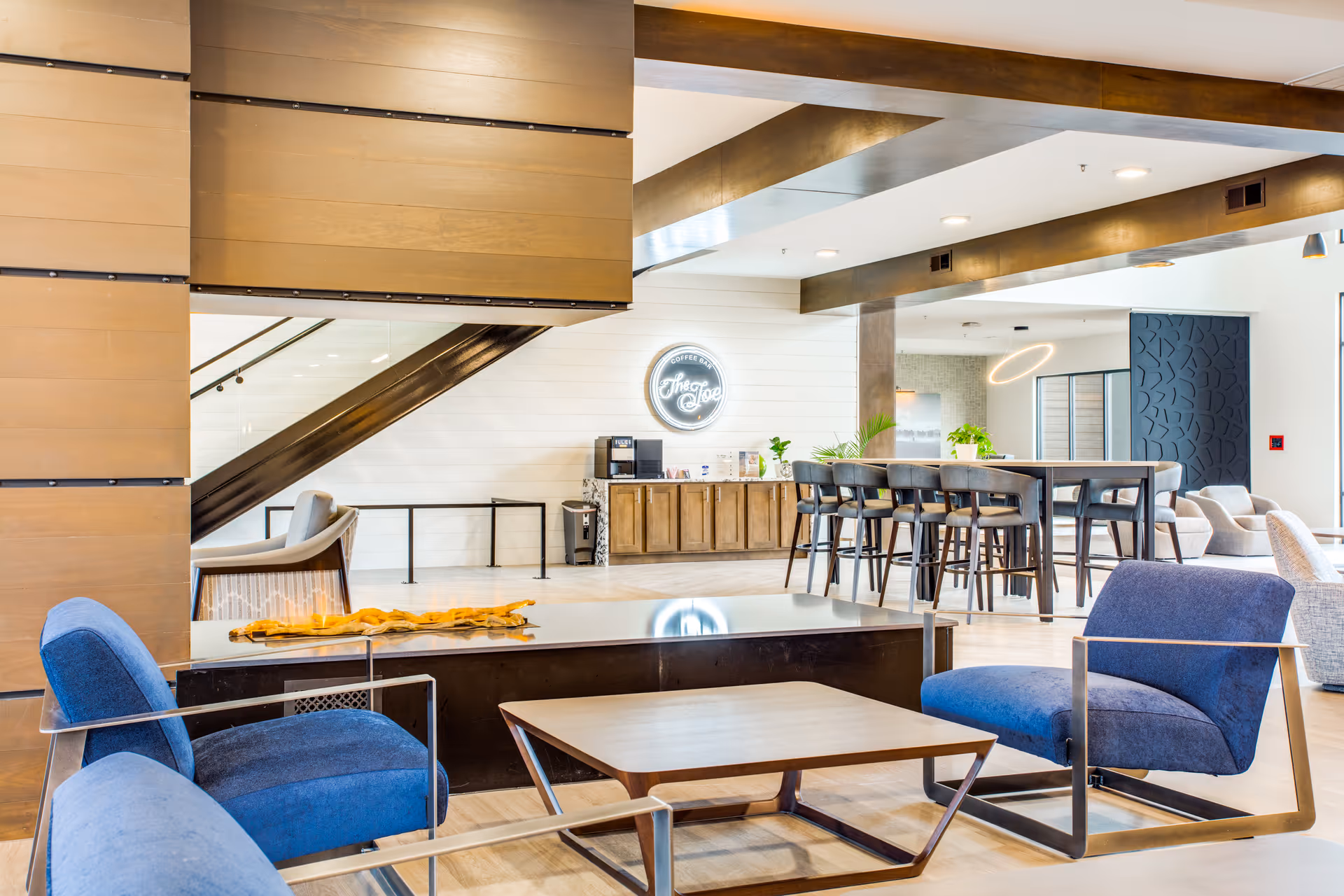 A modern and spacious lounge area with blue cushioned chairs and a wooden coffee table in the foreground. In the background, there is a coffee bar with a neon sign that reads 'The Joe', wooden cabinetry, and a high table with bar stools. The space features wooden beams on the ceiling and a staircase with glass railing.
