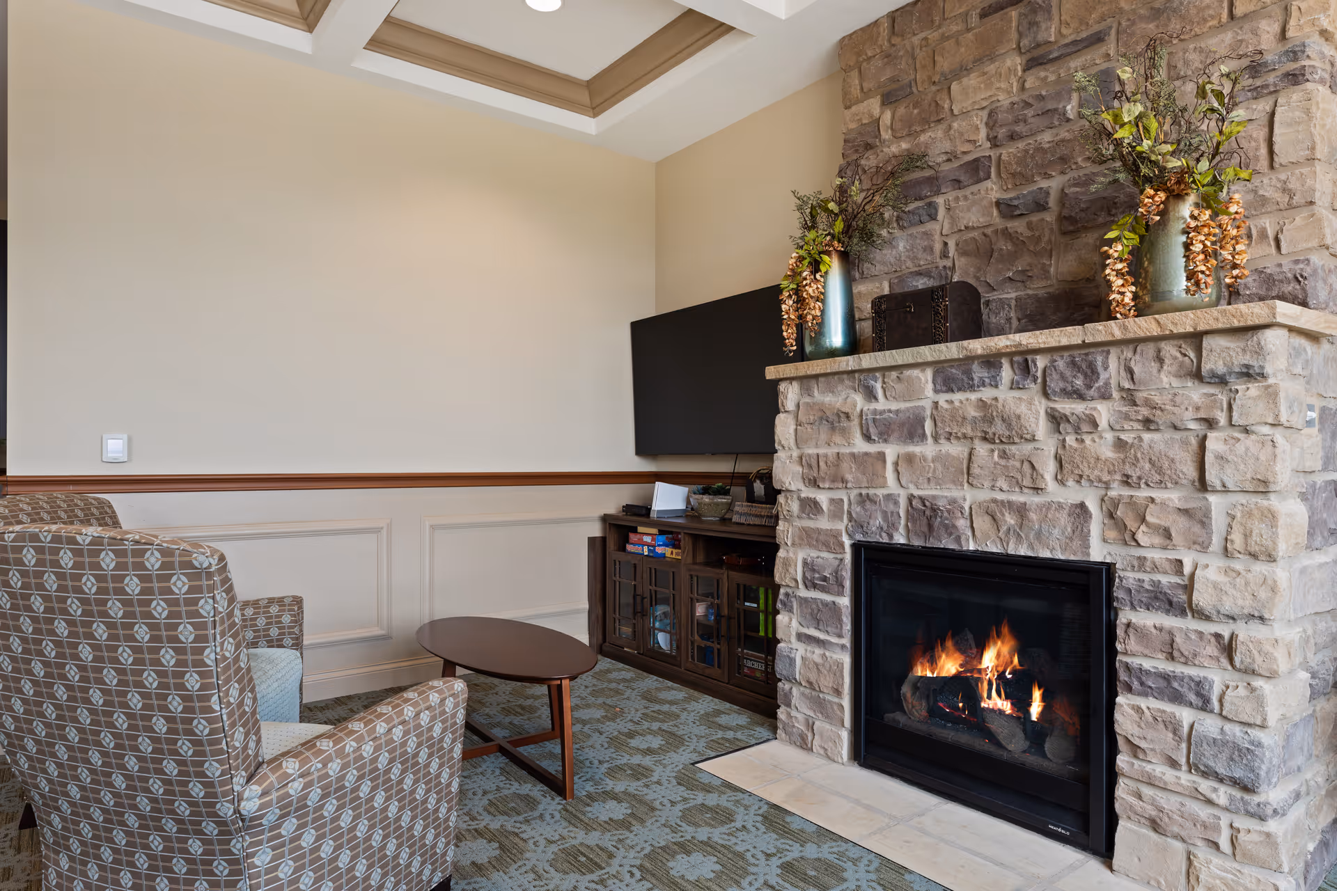 Cozy living room area with a stone fireplace burning firewood, two patterned armchairs, a small oval wooden coffee table, a flat-screen TV mounted on the wall, and a wooden cabinet with board games and decor. The room has beige walls with decorative molding and a patterned carpet.