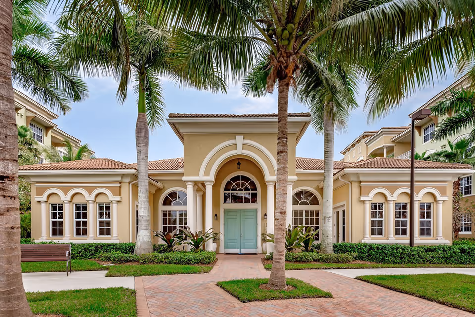 Mediterranean-style senior living clubhouse front with arched windows, a mint-green double-door entrance, and palm trees.