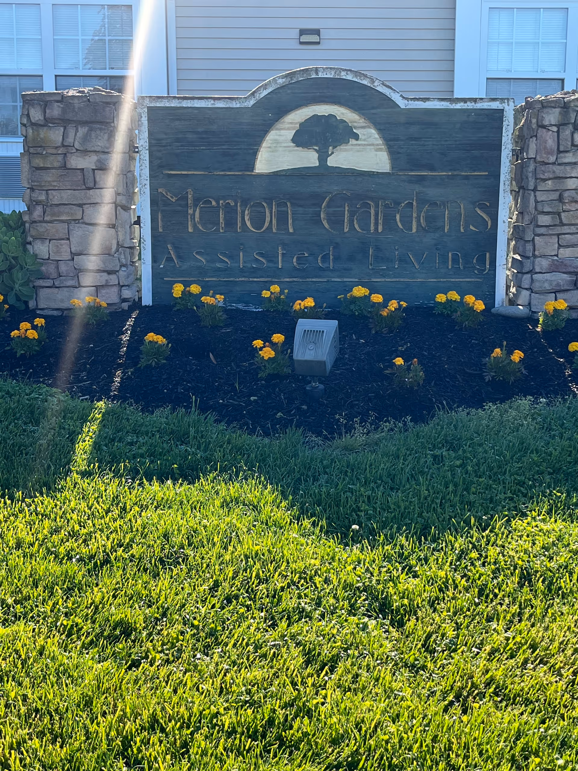 Wooden entrance sign reading "Merion Gardens Assisted Living" between stone pillars with yellow flowers and a grassy lawn.