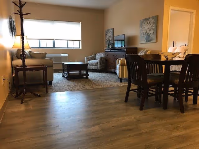 A cozy living area in a senior living facility featuring a wooden dining table with four chairs, a sitting area with a sofa, armchair, coffee table, and a TV on a wooden dresser. The room has wooden flooring, beige walls, and a window with a white blind letting in natural light.