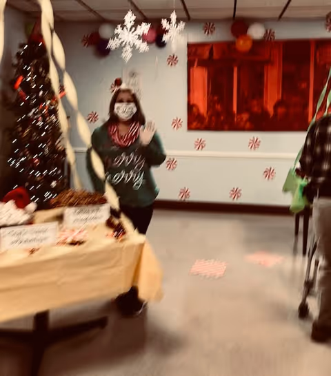 A decorated indoor room with a Christmas tree on the left, a table covered with a beige tablecloth displaying holiday treats, and a person wearing a green sweater and a face mask standing behind the table waving. The walls are decorated with red and white peppermint candy decals and hanging snowflake ornaments. Another person with a walker is partially visible on the right.