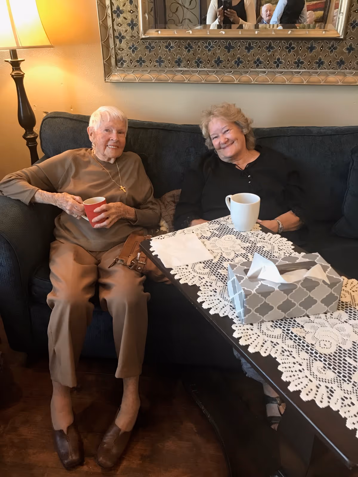 Two elderly women sitting on a dark blue couch in a cozy living room, each holding a cup. A table with a lace tablecloth and a tissue box is in front of them. A decorative mirror and a lamp are visible in the background.