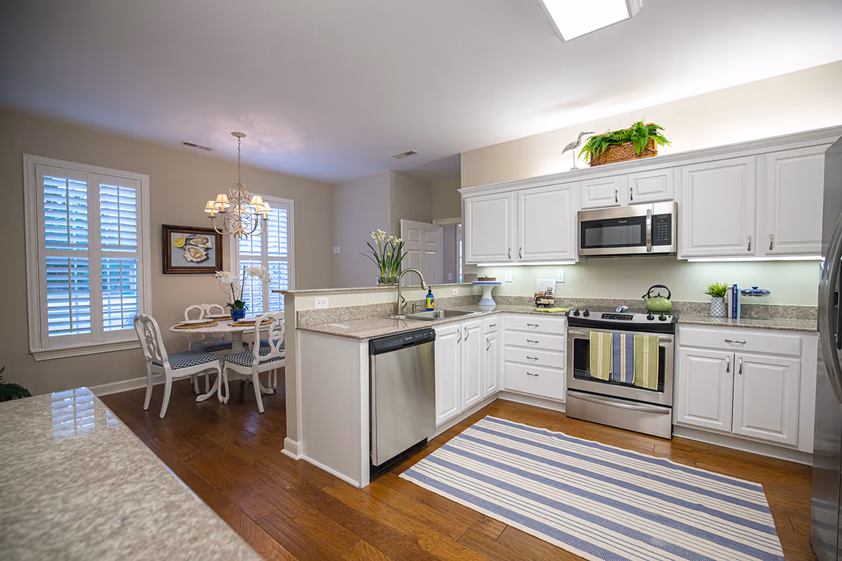 Bright kitchen with white cabinets, stainless steel appliances including a dishwasher, stove, and microwave. A striped rug is on the wooden floor. To the left, there is a small dining area with a round table and four chairs under a chandelier, next to two windows with white shutters.