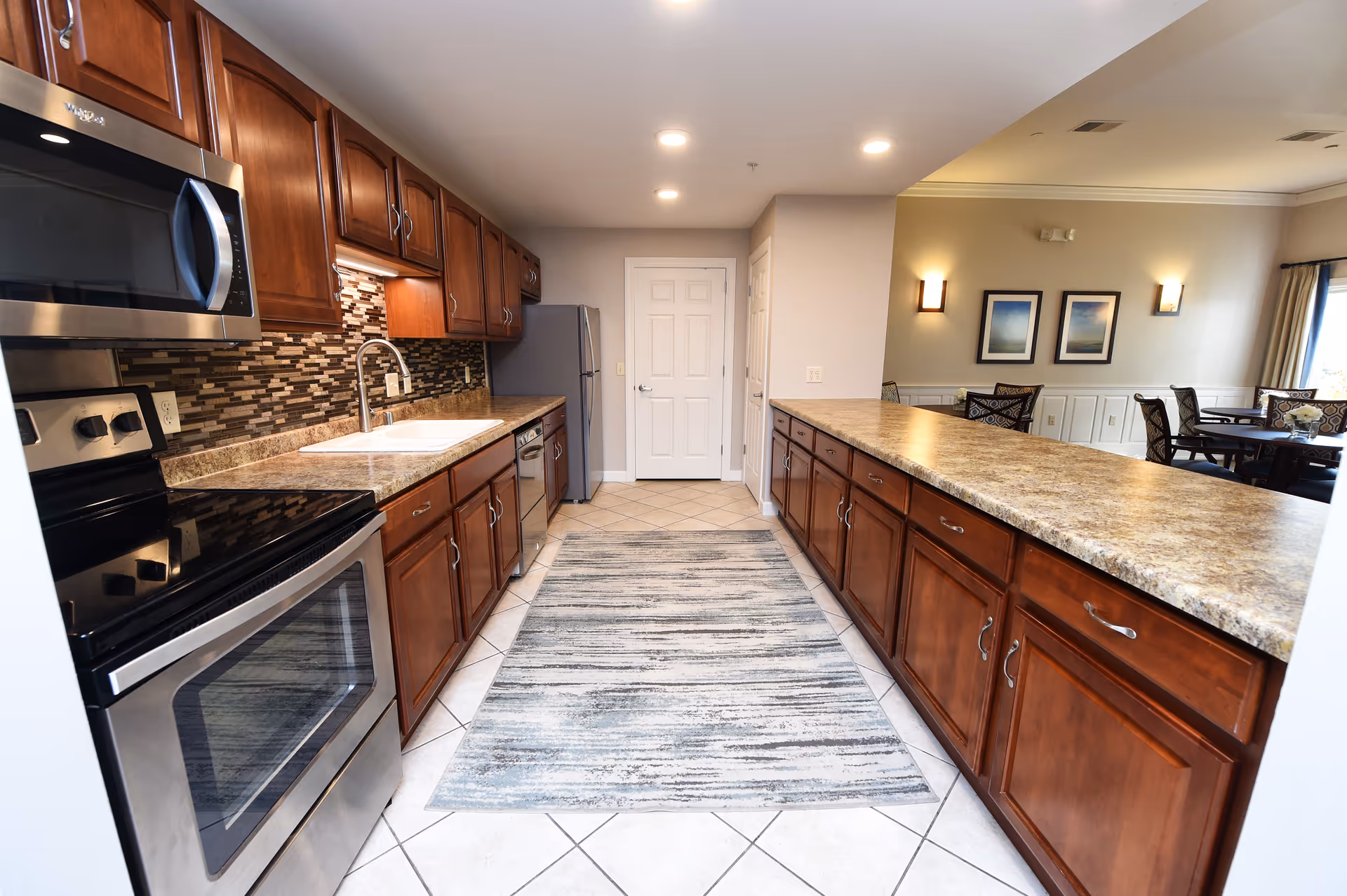 A modern kitchen with stainless steel appliances including a microwave, stove, and refrigerator. The kitchen features wooden cabinets, a tiled backsplash, a double sink, and a long countertop with a patterned rug on the tiled floor. In the background, there is a dining area with tables and chairs, wall sconces, and framed artwork on the walls.
