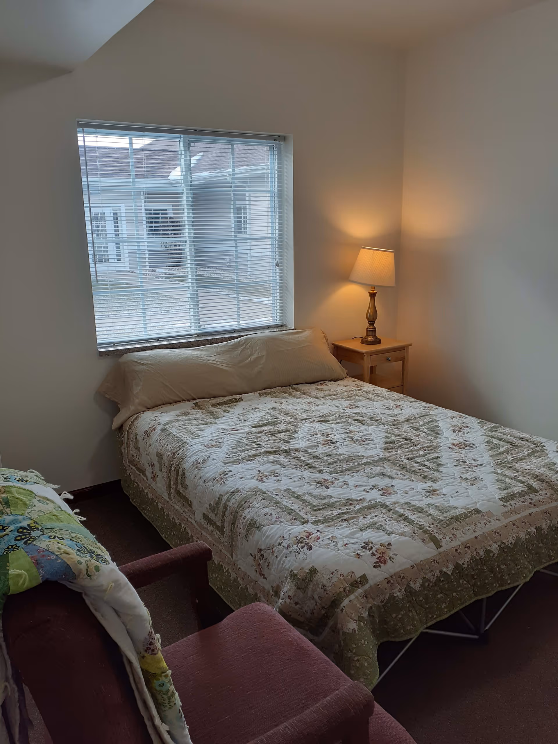 A small bedroom with a bed covered in a floral and green patterned quilt, a beige pillow, a wooden nightstand with a lit lamp, a window with blinds showing an outside view of a building, and a maroon armchair with a colorful quilt draped over it.