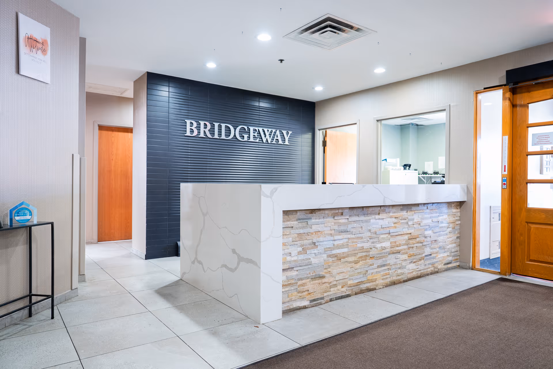 Reception area of Bridgeway of Bensenville Supportive Living with a modern white marble and stone front desk, a black tiled wall behind with the word 'BRIDGEWAY' in large silver letters, beige walls, tiled floor, and wooden doors.