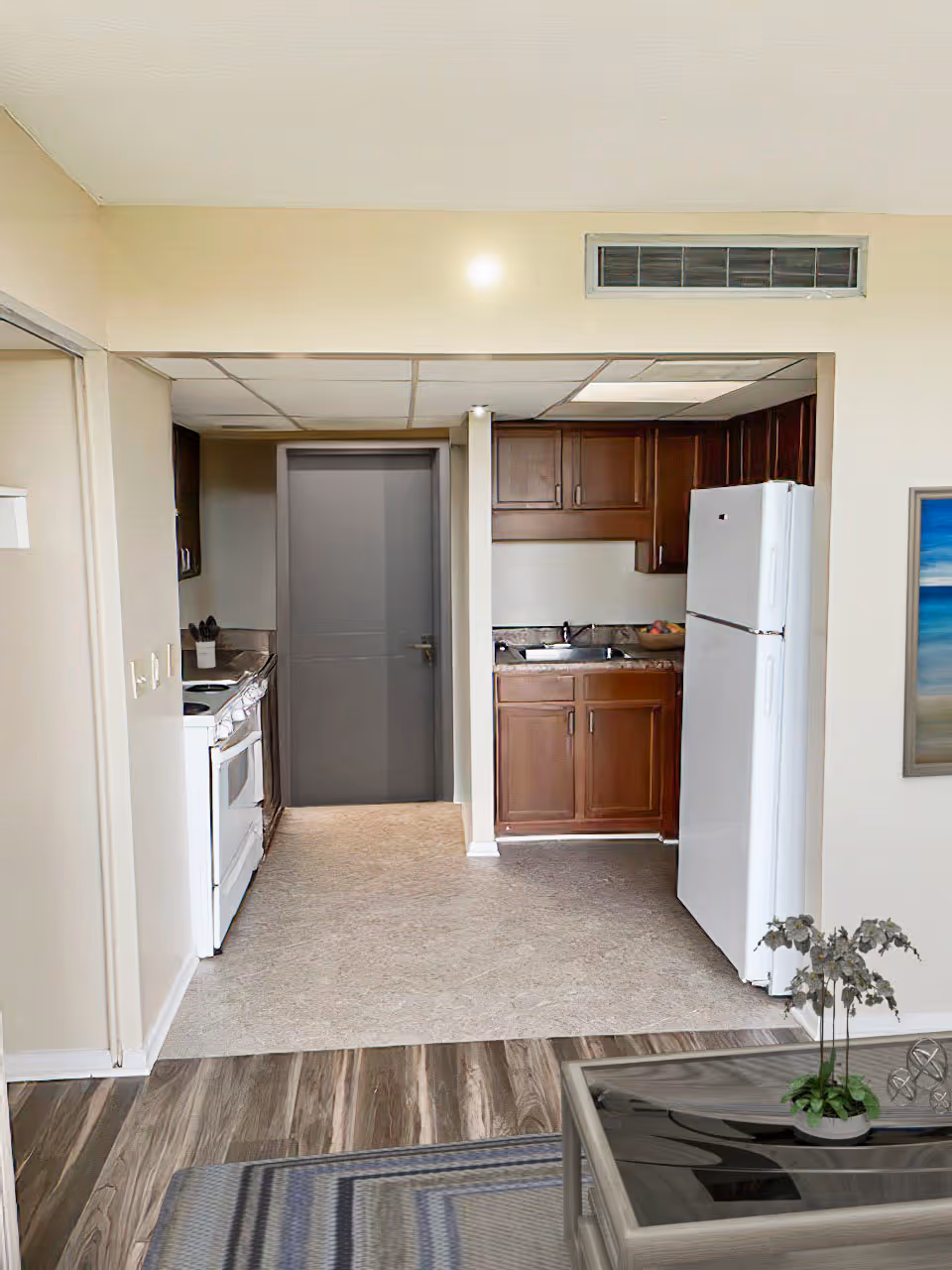 View of a small kitchen area with wooden cabinets, a white refrigerator, a white stove, and a sink. The kitchen floor is tiled, and the adjacent room has wood-patterned flooring with a glass coffee table and a small plant on it. A gray door is visible at the back of the kitchen, and a framed picture hangs on the wall to the right.