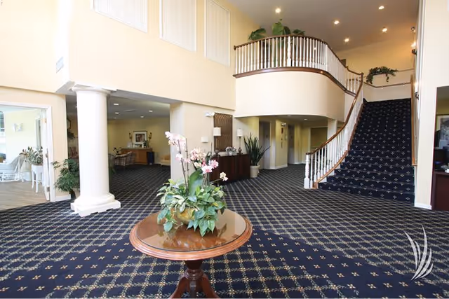 Spacious senior living community interior with patterned carpet, a round wooden table with a floral arrangement in the center, a curved staircase with white railings leading to an upper floor, and large white columns supporting the ceiling.