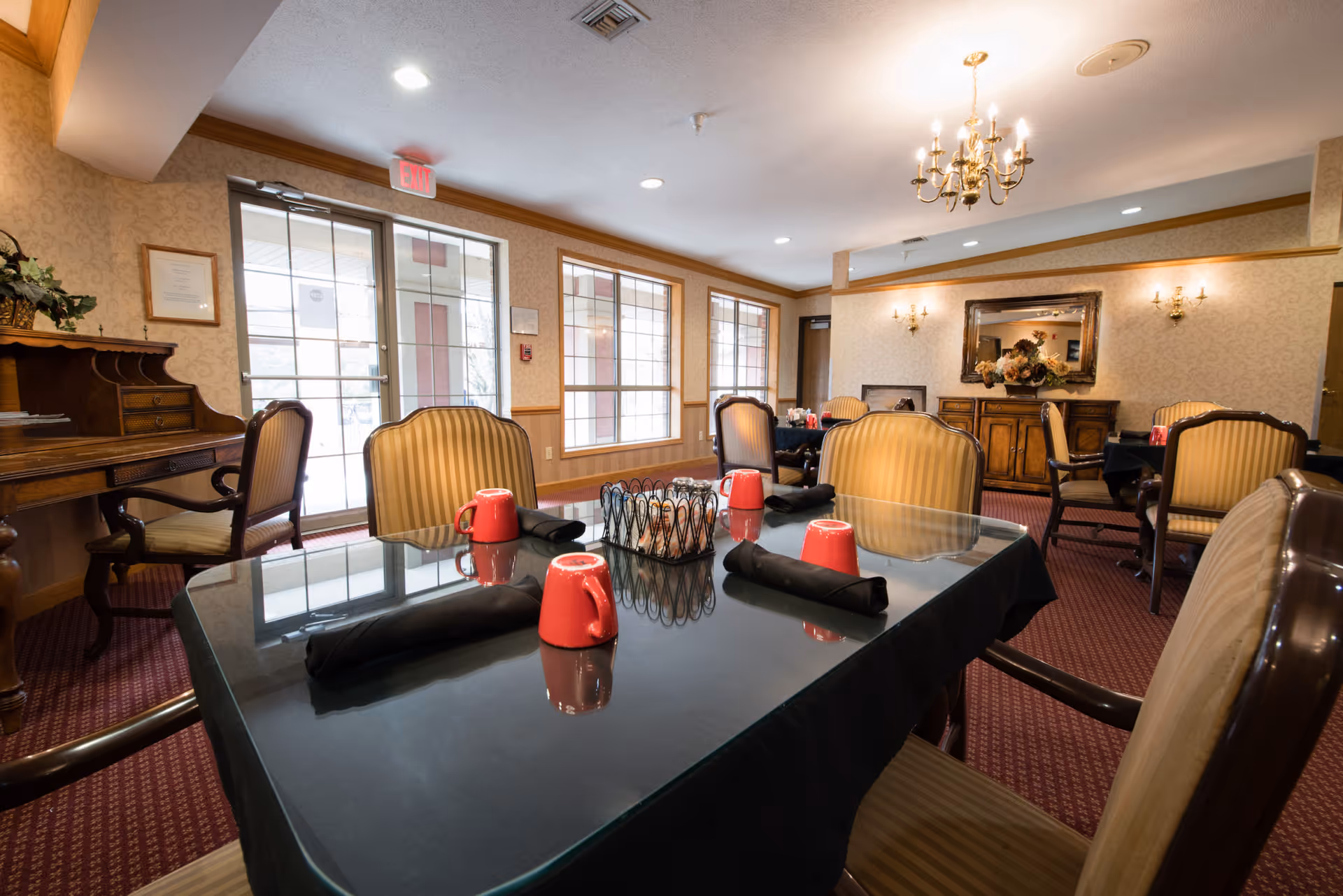 Dining room with multiple tables covered in black tablecloths, each set with red mugs and black napkins. The room has large windows letting in natural light, a chandelier, wall sconces, a decorative mirror, and a wooden sideboard with a floral arrangement. There is also a wooden desk and chairs along one wall.