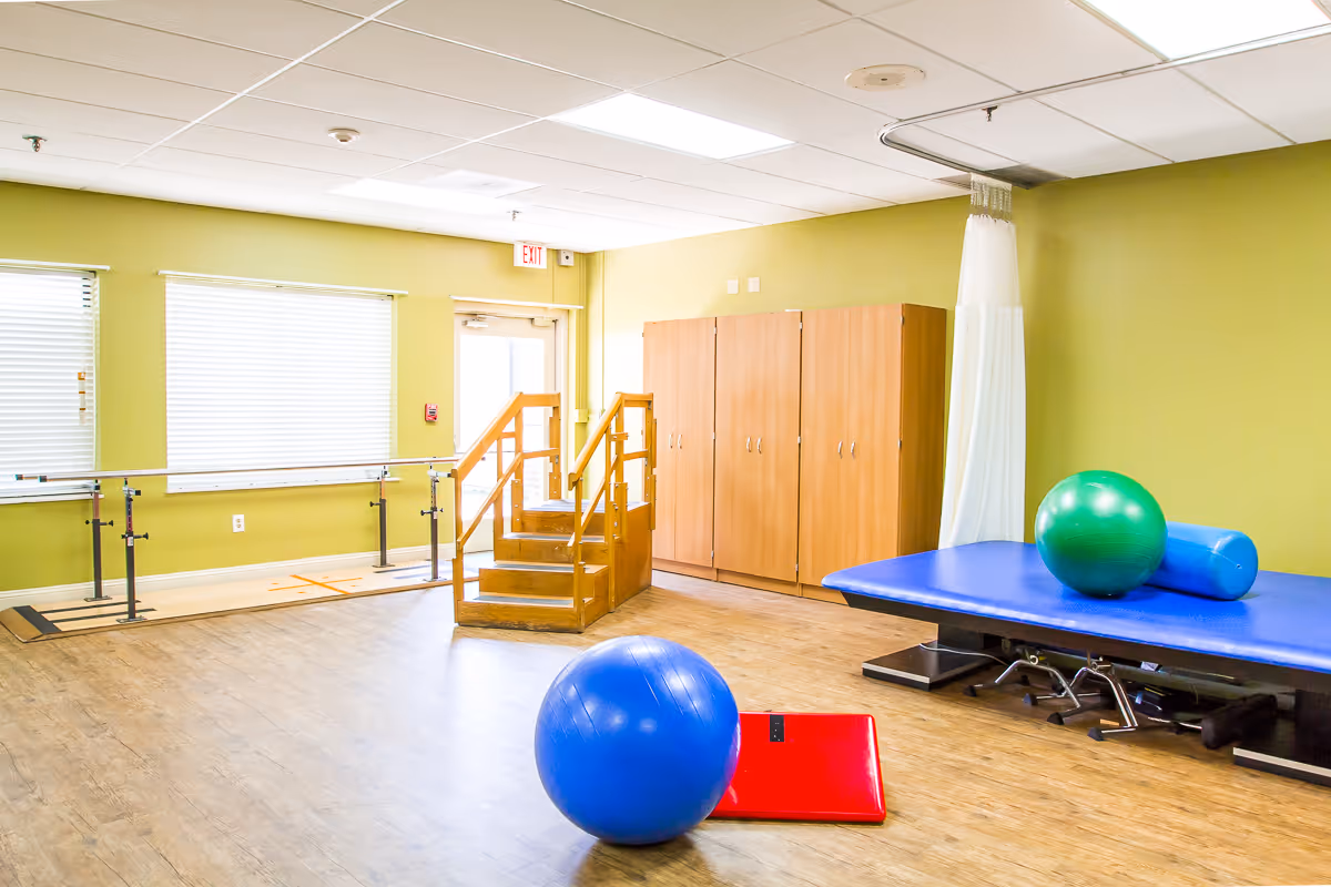 A physical therapy room with wooden stairs and handrails for rehabilitation, a blue therapy table with exercise balls on top, a red mat on the floor, and wooden cabinets against a green wall. The room has large windows with blinds and a wooden floor.