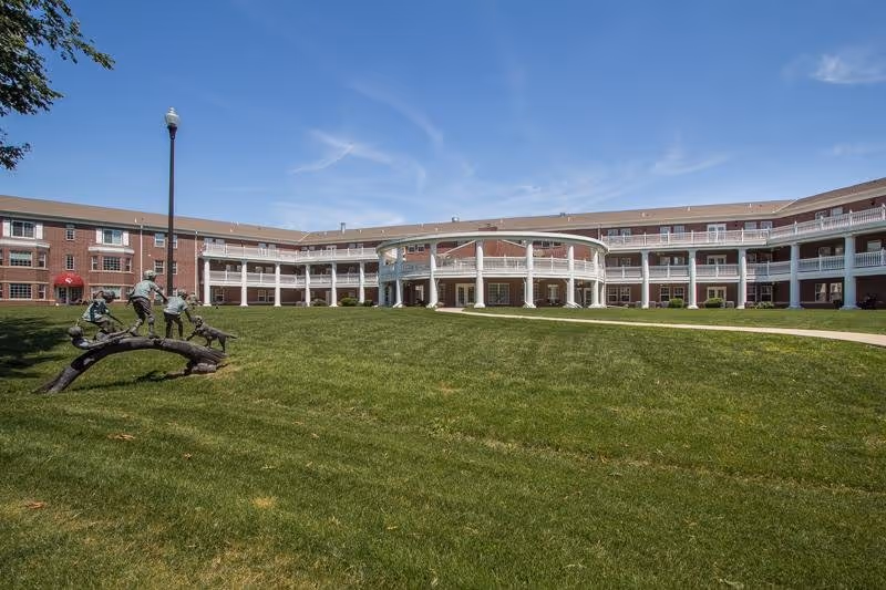 Wide view of Carnegie Village Senior Living Community building with a large green lawn in front, a curved two-story brick building with white columns and balconies, and a sculpture of children playing on a curved branch near a lamppost under a clear blue sky.