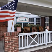 Front porch area of a senior living facility with a white railing, brick pillars, rocking chairs, an American flag, and a sign that reads 'Aspen'.