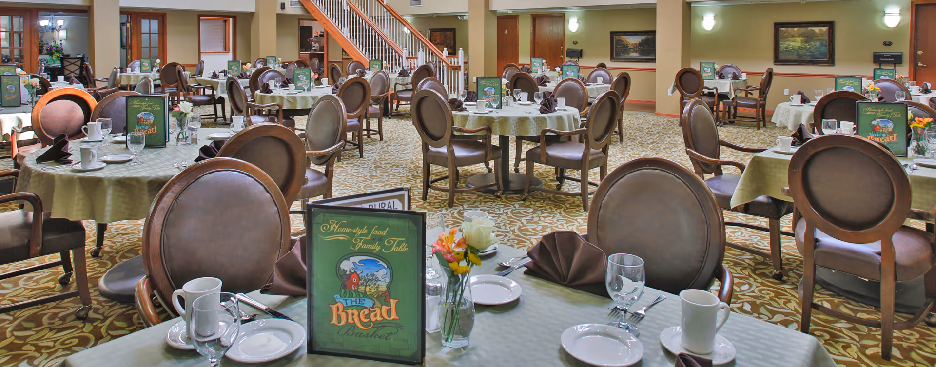 A spacious dining room with multiple round tables covered with light green tablecloths, each set with white plates, cups, glasses, silverware, and brown folded napkins. Menus titled 'The Bread Basket' and small flower vases are placed on the tables. The room features patterned carpet, wooden chairs with cushioned seats and backs, wall paintings, and a staircase in the background.