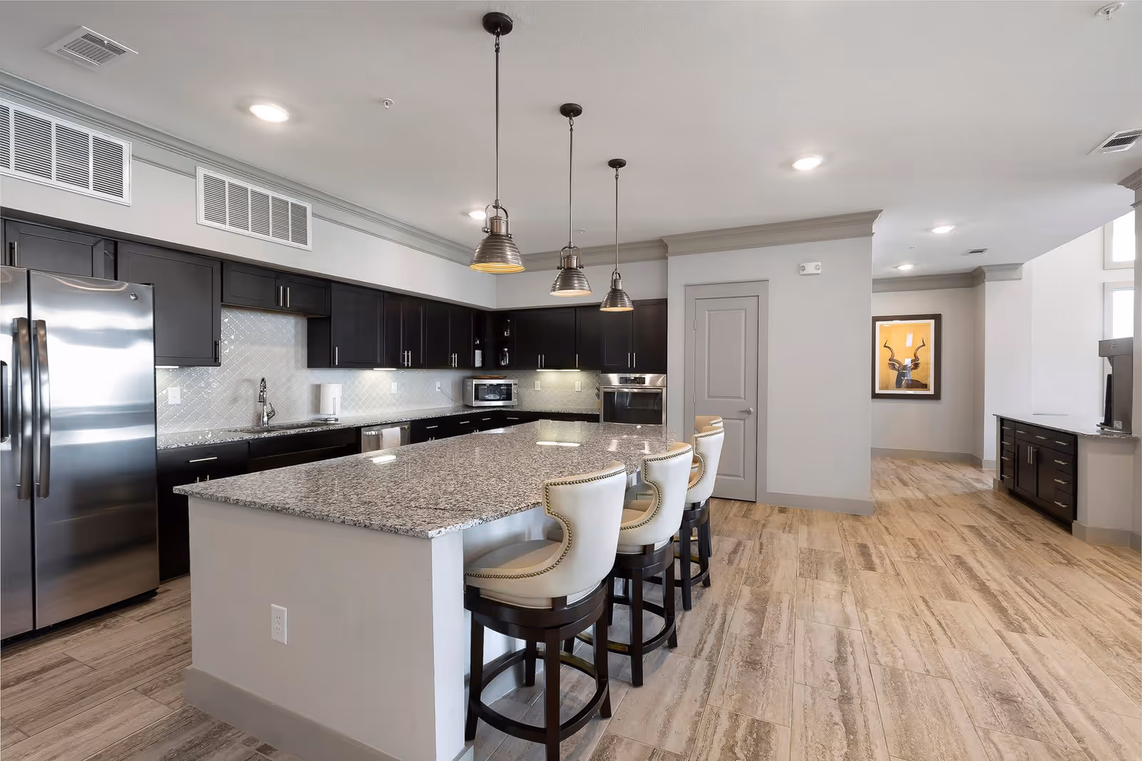 A modern kitchen with a large granite island countertop surrounded by four white cushioned bar stools with dark wooden legs. The kitchen features dark wood cabinets, a stainless steel refrigerator, built-in oven, microwave, and under-cabinet lighting. Three industrial-style pendant lights hang above the island. The floor is covered with light wood-patterned tiles, and a framed artwork is visible on the wall in the background.
