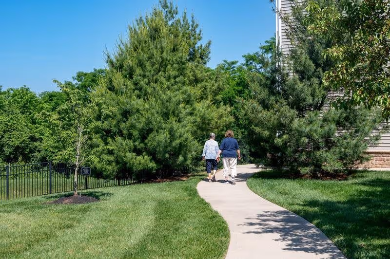 Two elderly women walking on a curved concrete pathway surrounded by green grass and trees on a sunny day near a building.