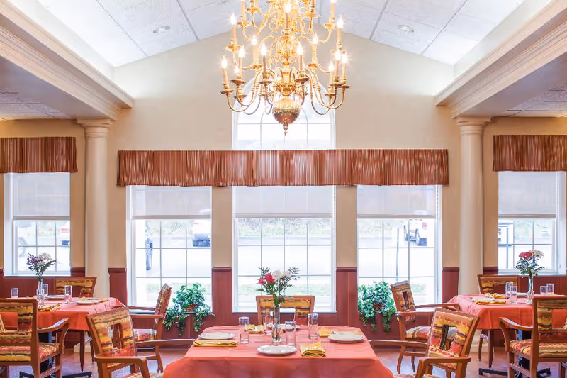A bright dining room with several tables covered in red tablecloths, each set with plates, glasses, and yellow napkins. Wooden chairs with patterned cushions surround the tables. Large windows with striped valances let in natural light, and a large ornate chandelier hangs from the ceiling. Green plants are placed near the windows.