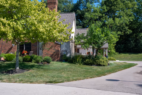 Exterior view of a senior living facility with a brick building partially visible, surrounded by green trees, shrubs, and a well-maintained lawn under a clear blue sky.