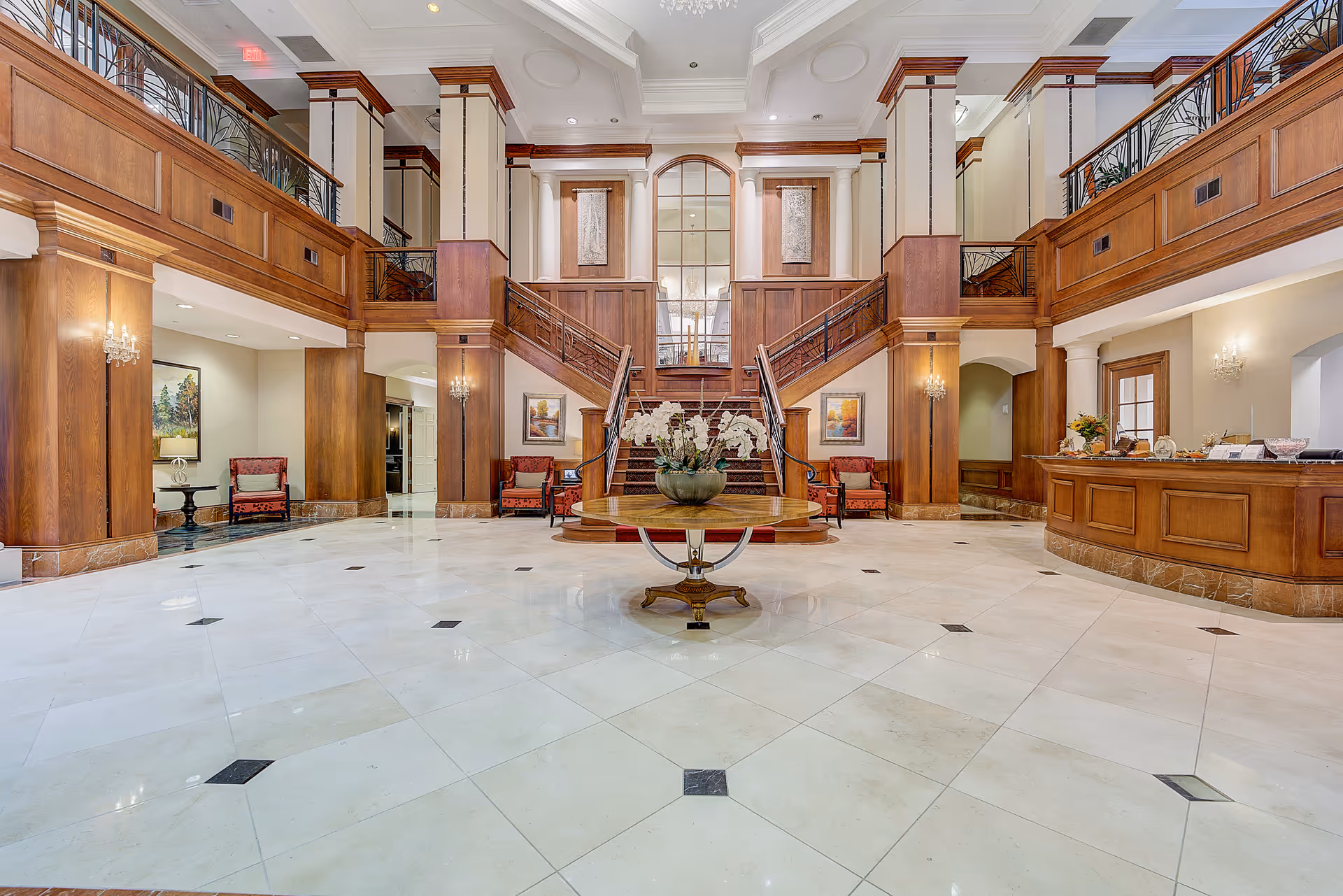 Spacious and elegant lobby area with a central round table holding a floral arrangement, polished marble floor with black diamond accents, wooden paneling and columns, a grand staircase with wrought iron railings leading to an upper level, comfortable seating areas with red upholstered chairs, wall sconces, and framed artwork.