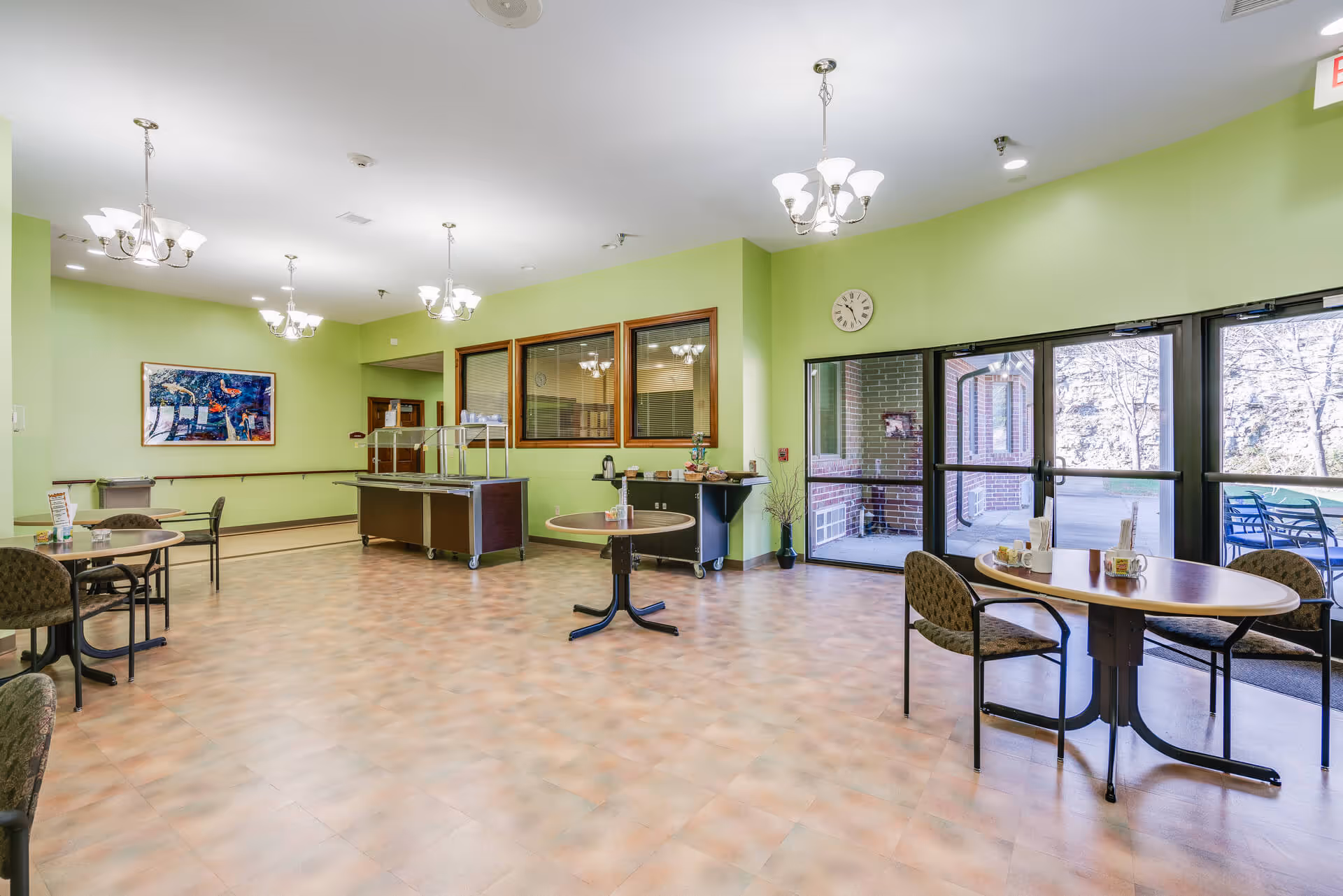 A bright dining area in a senior living facility with light green walls, several round tables with chairs, a serving station, and large glass doors leading outside to a patio with outdoor seating. The room is well-lit with multiple ceiling chandeliers and has a wall clock and a colorful painting on the wall.