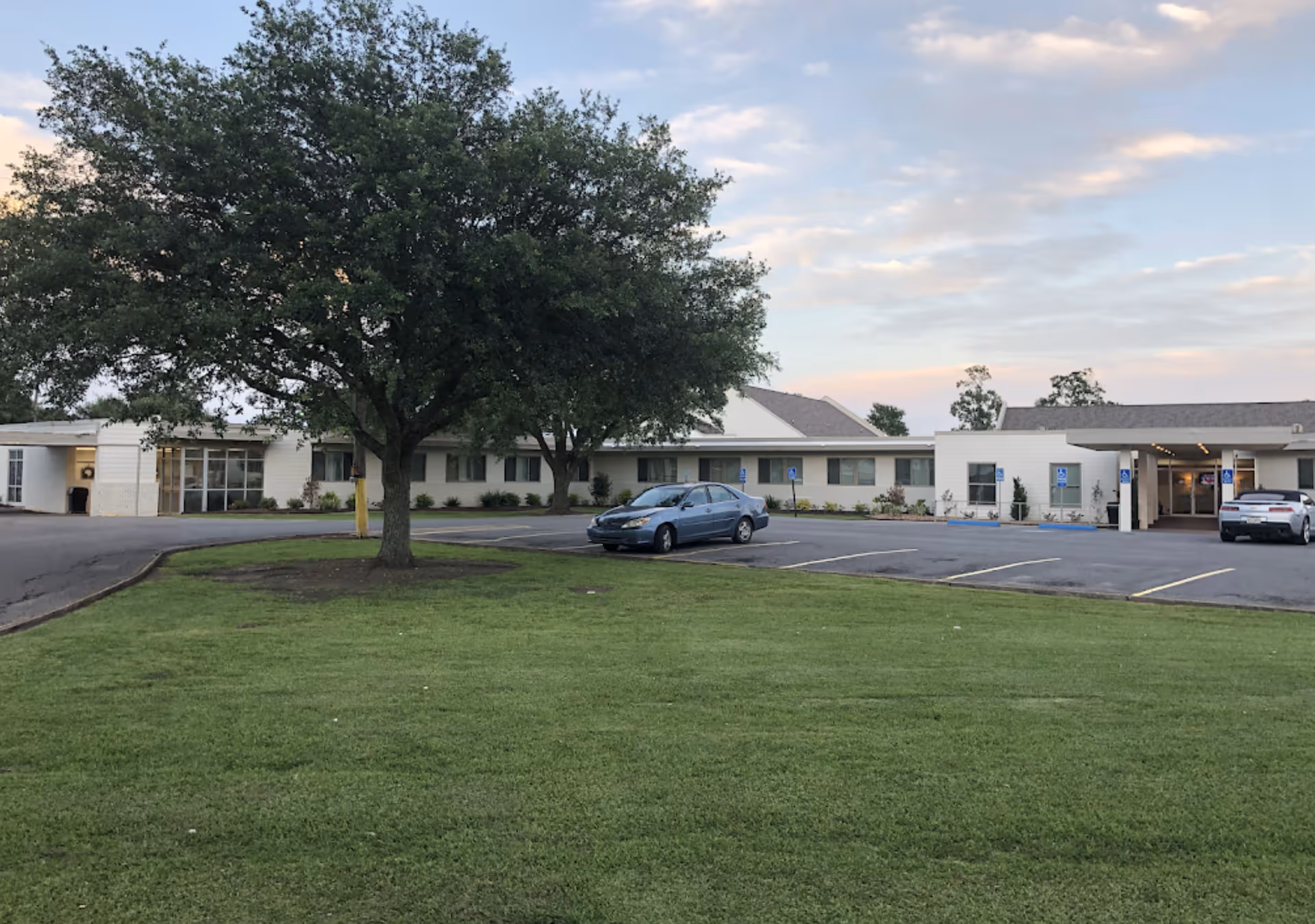 Low single-story senior living building with a parking lot, a large tree, and a grassy lawn under a partly cloudy sky.
