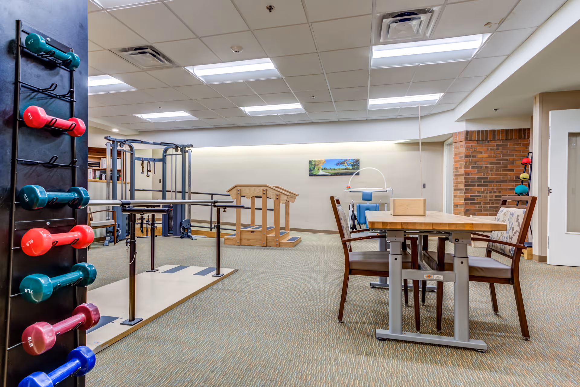 Interior view of a physical rehabilitation center room with exercise equipment including parallel bars, a wooden step platform, and a rack of colorful dumbbells. There is a table with chairs on the right side and a brick accent wall in the background.